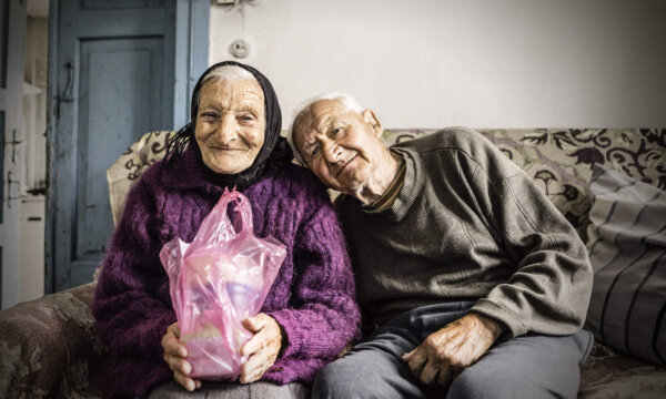 Ein älterer Mann legt seinen Kopf an die Schulter einer älteren Dame. Diese hält eine Tüte mit Hilfsgütern in der Hand.
