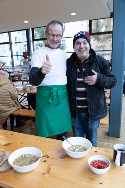 Mitarbeiter der Suppenküche der Franziskaner und ein Bedürftiger stehen Arm in Arm vor einem gedeckten Tisch und geben das "Daumen Hoch"-Zeichen