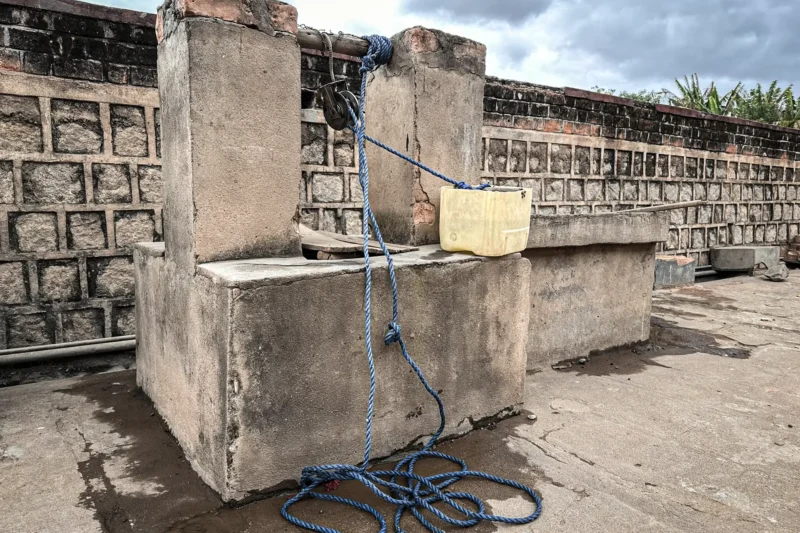 Ein traditioneller gemauerter Brunnen mit einem blauen Seil und einem gelben Plastikkanister als Wasserbehälter, daneben eine niedrige Mauer aus Steinblöcken, unter einem bewölkten Himmel.