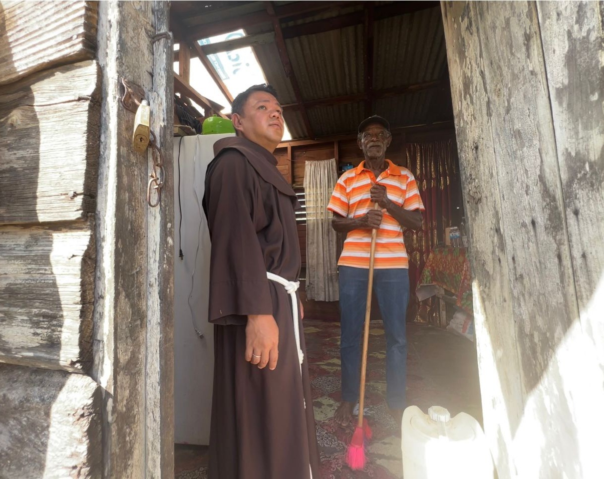 Franziskaner in Habit steht neben einem Mann mit Besen in der Hand in einem Holzhaus mit beschädigtem Blechdach auf Jamaika.