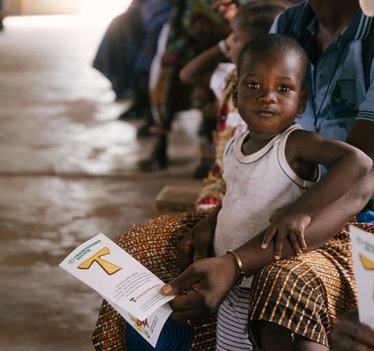 Kleines Kind sitzt auf dem Schoß einer Frau in einer Warteschlange und schaut in die Kamera, während die Frau einen Flyer von Franziskaner Helfen in der Hand hält.