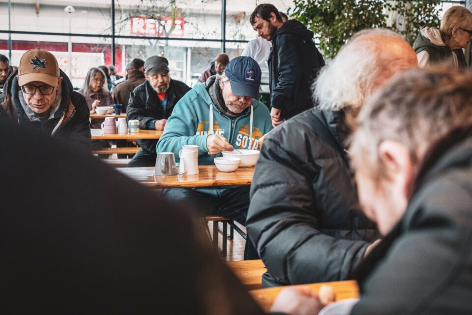 Menschen sitzen an Tischen in einer Suppenküche und essen gemeinsam eine warme Mahlzeit.