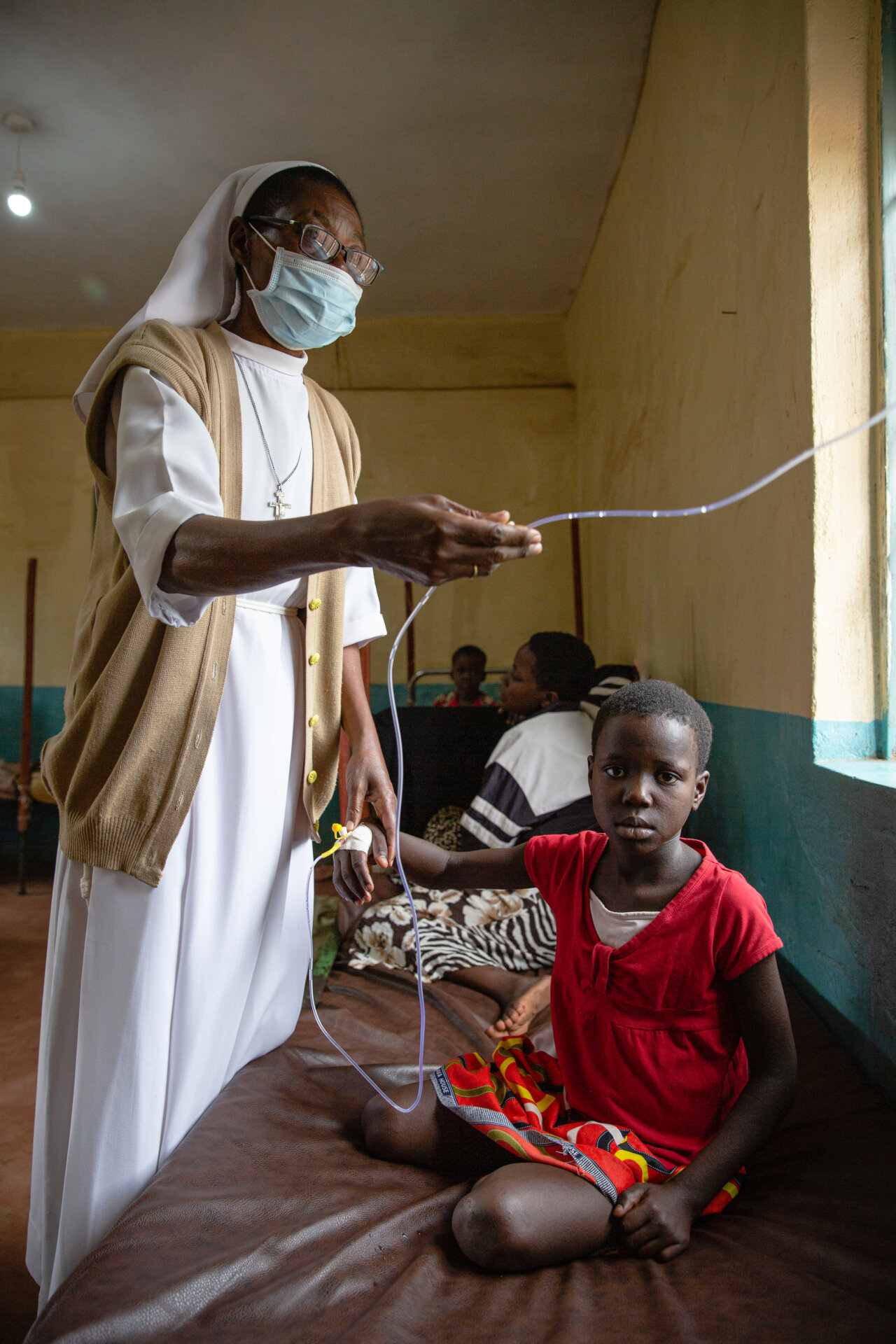 Eine Franziskanerin im weißen Habit steht an dem Krankenbett und überprüft die Flüssigkeit im Venenkathether mit der einen Hand. Mit der anderen hält sie die Hand des kleinen Mädchens, das auf dem Bett sitzt.