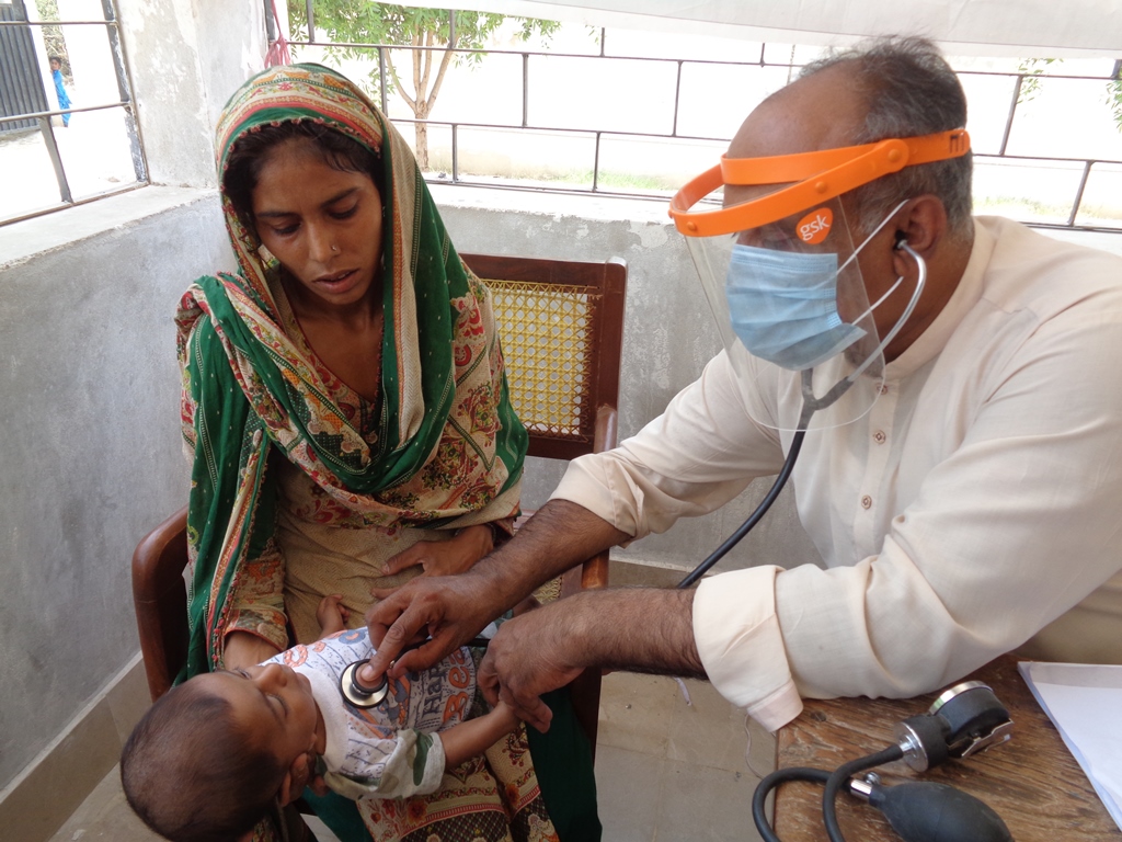 Ein pakistanischer Arzt untersucht ein Baby, dass auf dem Schoß seiner pakistanischen Mutter liegt. Die Mutter trägt ist traditionell gekleidet.