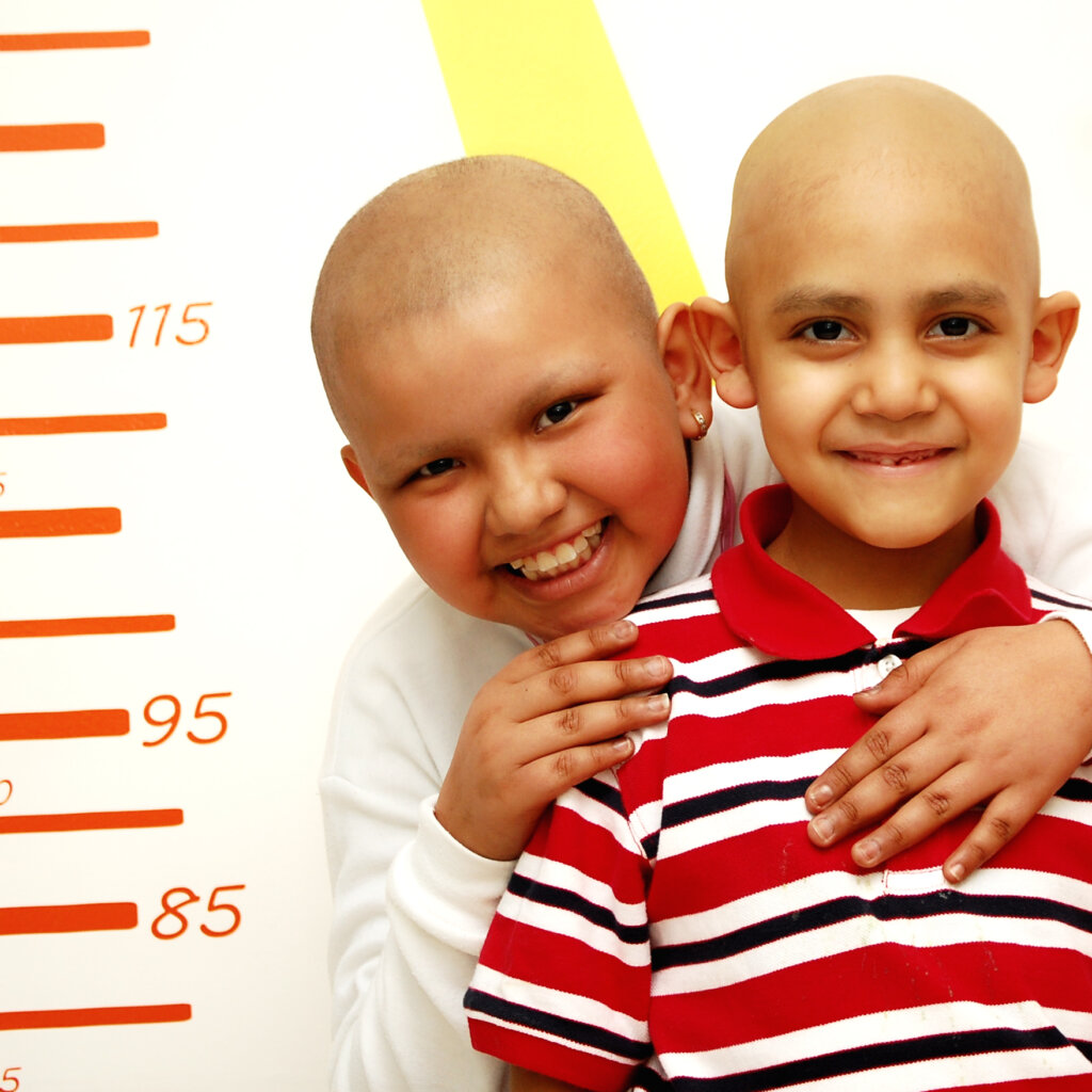 Zwei kahlköpfige Jungen, die eng beieinander stehen und in die Kamera lächeln. Der eine Junge umarmt den anderen von hinten; beide wirken freundlich und zugewandt. Links ist ein farbiger Messbalken zu sehen.