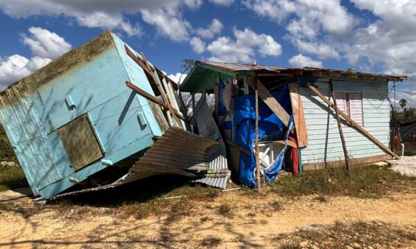 Zwei kleine Häuser stehen nebeneinander; eines ist zur Seite gekippt und stark beschädigt. Wellblech und Holzteile sind verbogen, der Boden ist sandig, darüber ein blauer Himmel mit Wolken.