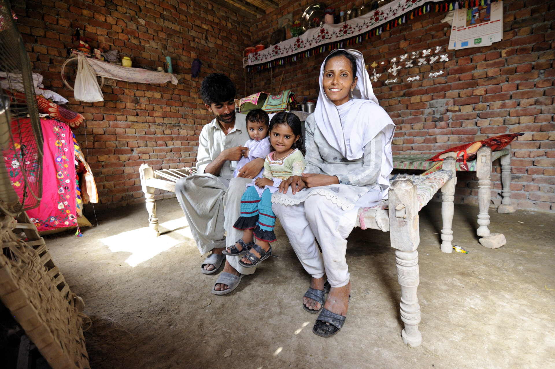 Pakistanische arme Familie in einem Haus aus Ziegeln. Die vierköpfige Familie sitzt in einer Reihe auf einem Bett. Die Eltern außen, die kleinen Kinder (Mädc hen und Junge) innen.