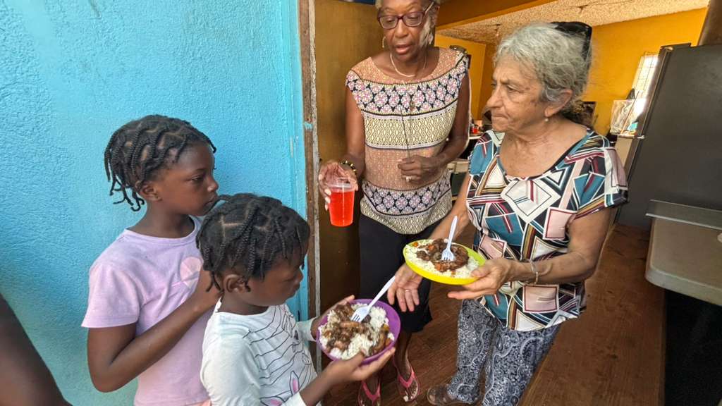 Eine Frau verteilt Essen an Kinder an der Tür eines Hauses. Die Kinder halten Teller in den Händen und warten.