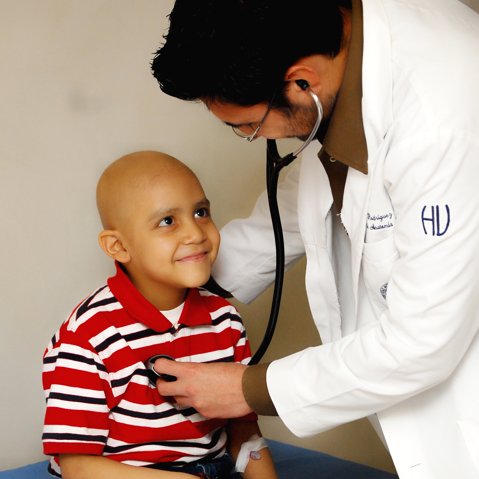 Ein Arzt in weißem Kittel untersucht mit einem Stethoskop einen kleinen Jungen im rot-weiß gestreiften Polohemd. Der Junge schaut den Arzt an und lächelt. Er ist leukämiekrank.