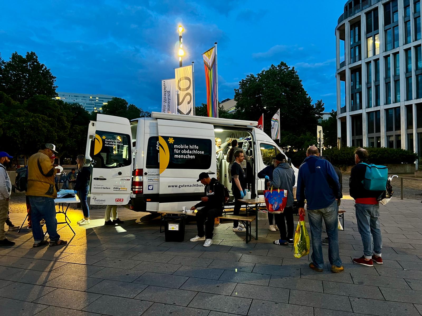 Ein Kleinbus steht abends auf einer Straße. Mehrere Menschen stehen davor im Gespräch