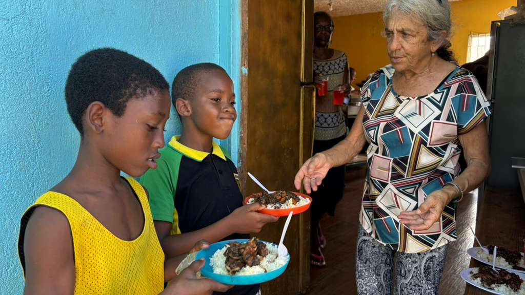 Eine ältere Frau reicht Kindern Teller mit Essen an der Tür eines Hauses. Die Kinder stehen dicht beieinander vor der Haustür und nehmen das Essen entgegen.