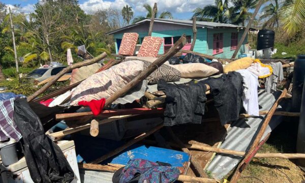 Vor einem kleinen Haus liegt ein großer Trümmerhaufen aus Holz und Wellblech. Zwischen den Trümmern hängen schwarze Planen, im Vordergrund sind verstreute Kleidungsstücke zu sehen. Im Hintergrund stehen Bäume unter blauem Himmel.