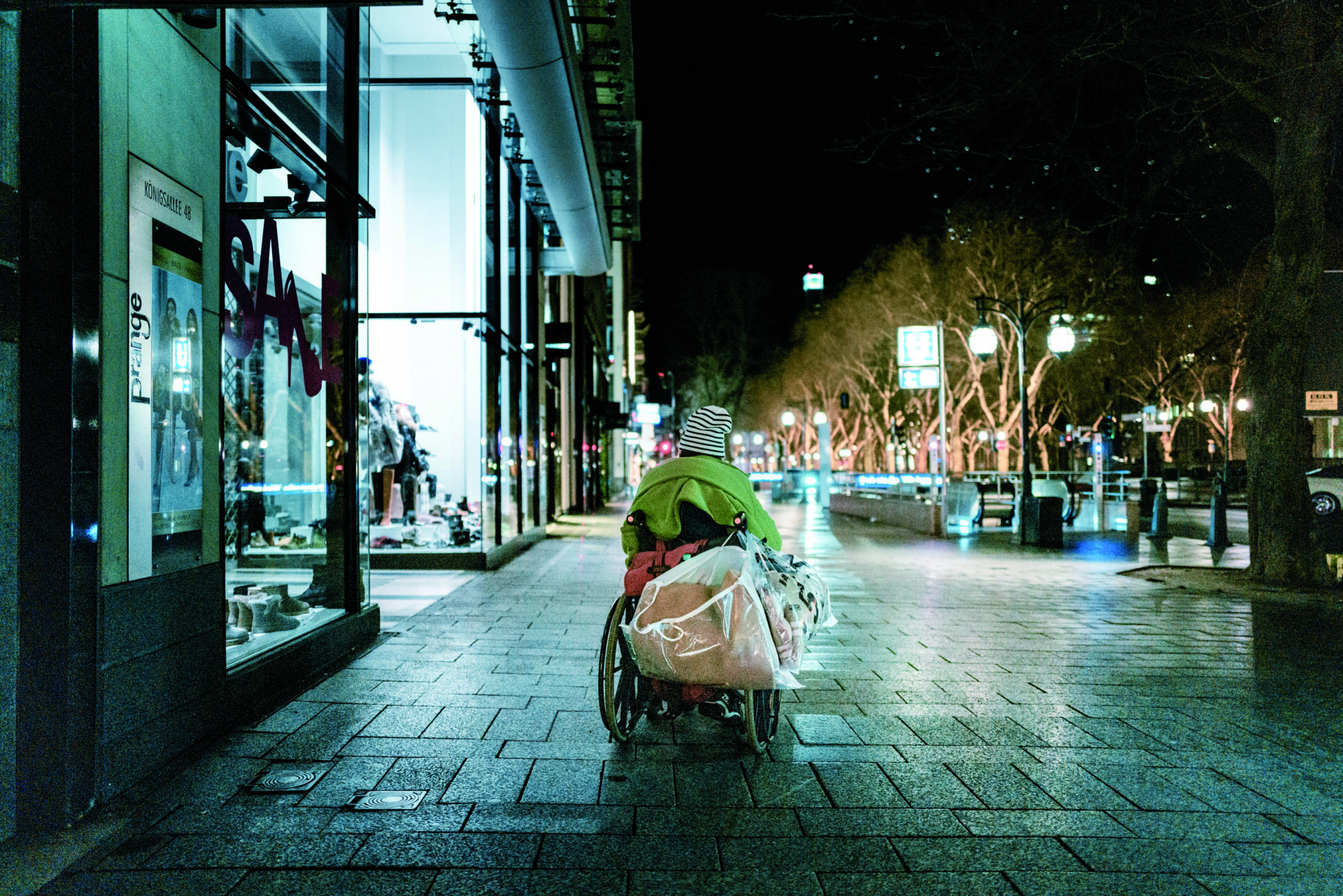 Eine Person ist nachts von hinten in einem Rollstuhl zu sehen. Sie befindet sich auf einer nassen Einkaufsstraße vor einem Gebäude mit großen beleuchteten Schaufenstern.