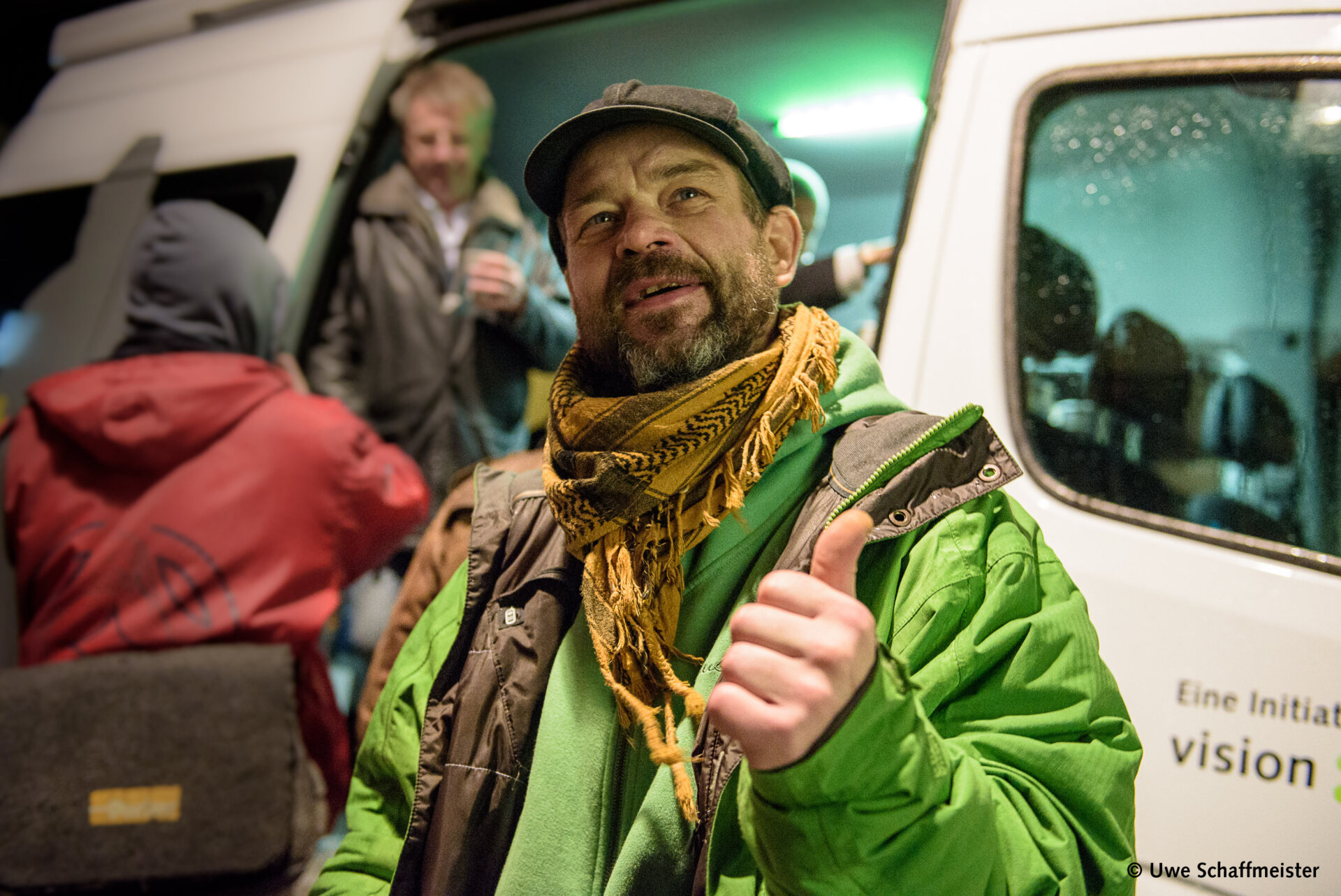 Ein Mann mit Mütze und Schal steht vor einem Bus und zeigt den Daumen nach oben. Im Hintergrund sind weitere Personen und die offene Bustür zu sehen.