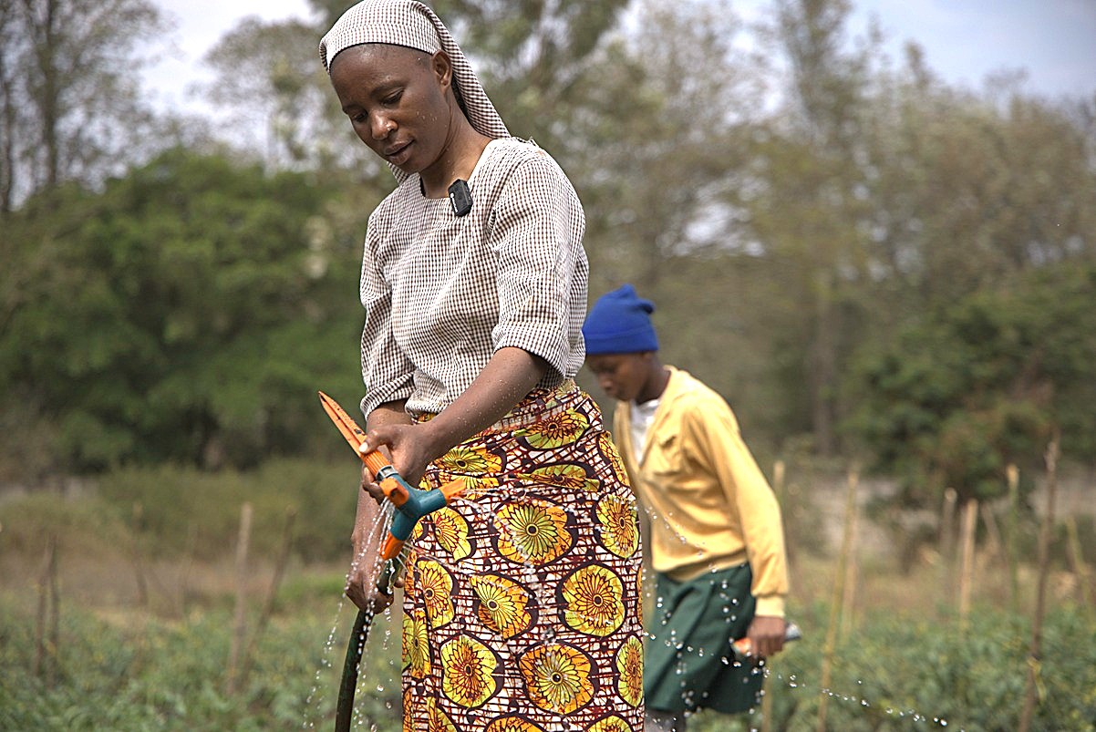 Eine Frau bewässert mit einem Schlauch ein Feld in Tansania, während im Hintergrund eine weitere Person bei der Gartenarbeit hilft.
