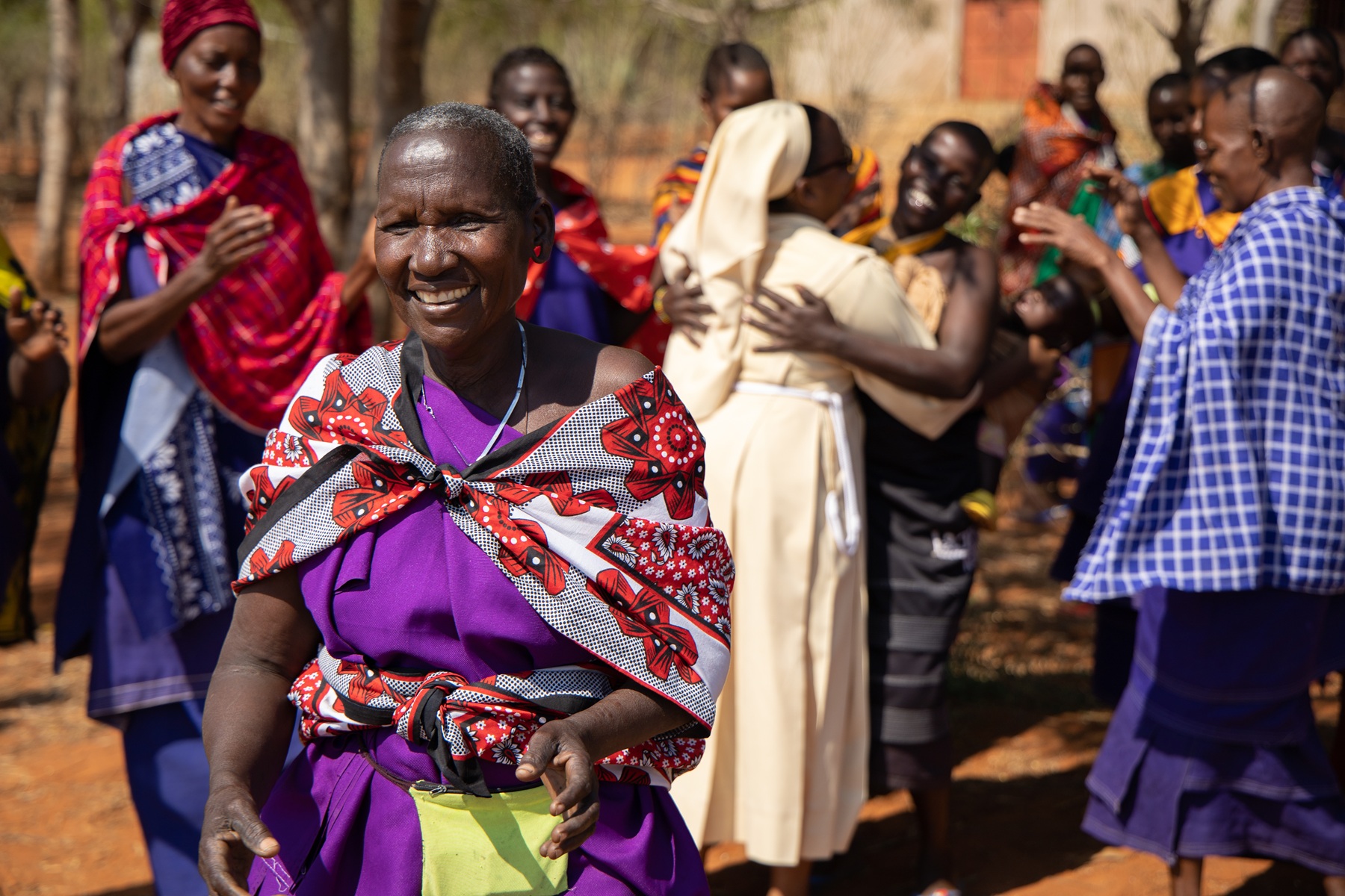 Eine Frau lacht im Vordergrund, während im Hintergrund Menschen eine Franziskanerschwester herzlich begrüßen und umarmen.