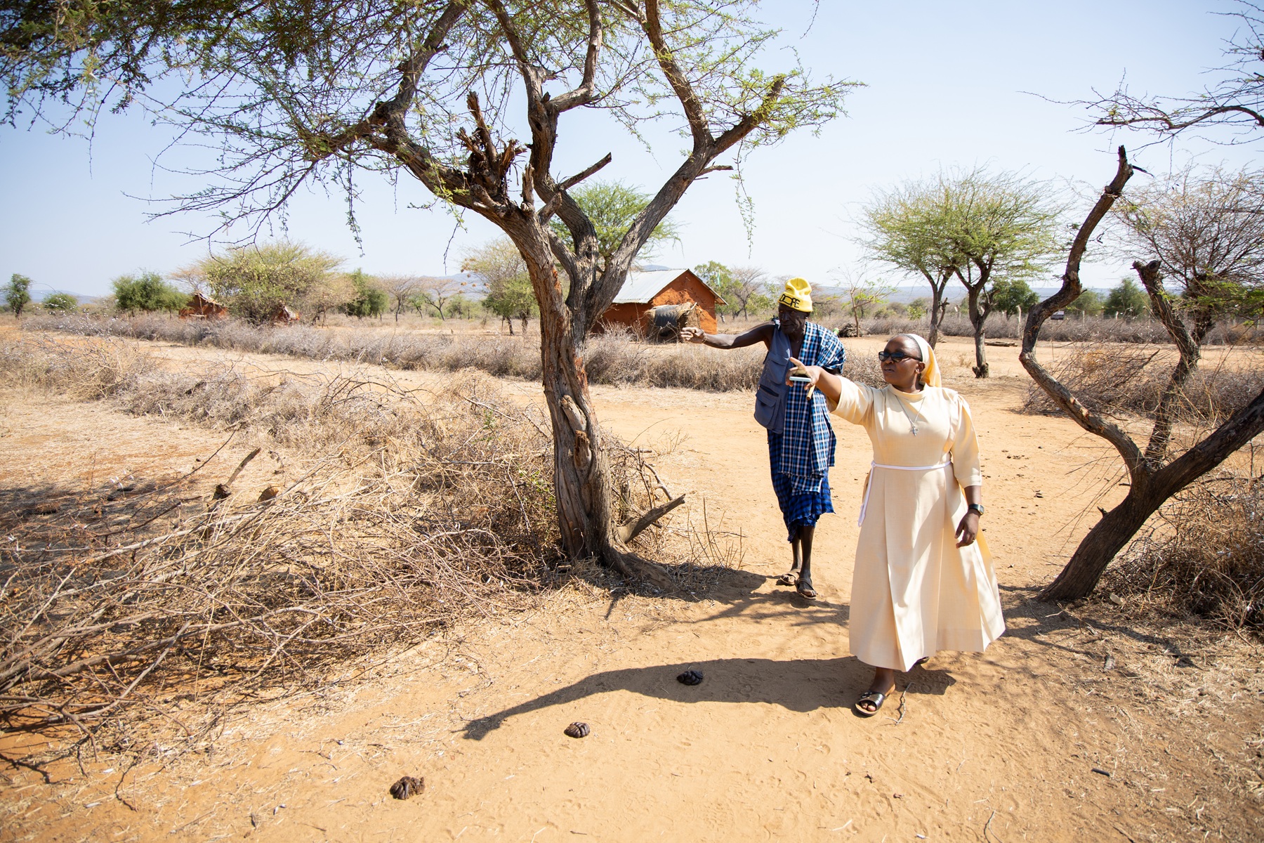 Eine Franziskanerschwester und ein Mann gehen durch eine trockene Landschaft und zeigen in die Ferne, umgeben von Bäumen und einfachen Häusern.