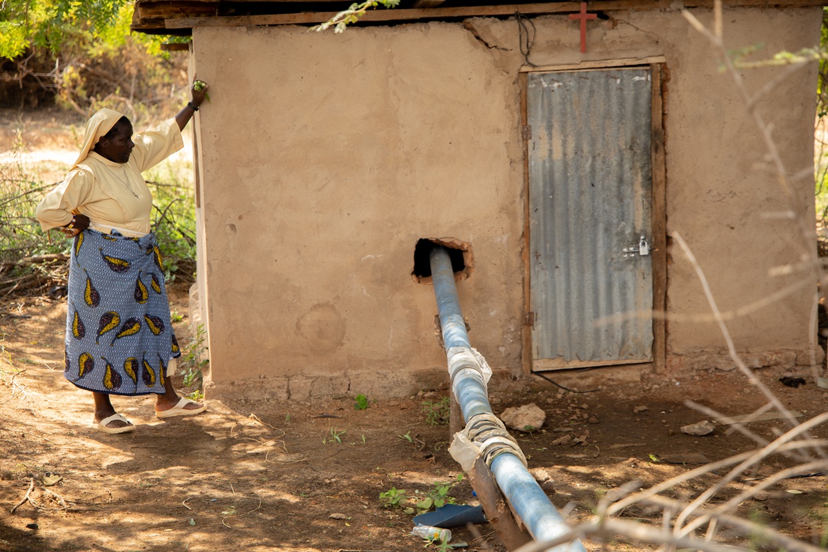 Eine Franziskanerschwester steht neben einem kleinen Haus mit Wasserleitung und betrachtet die Installation an der Außenwand
