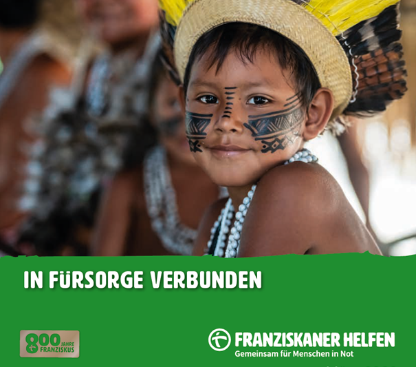 Ein indigenes Kind mit bemaltem Gesicht und traditionellem Federschmuck blickt in die Kamera. Es trägt eine Kette aus Perlen; im Hintergrund sind weitere Personen unscharf zu erkennen.