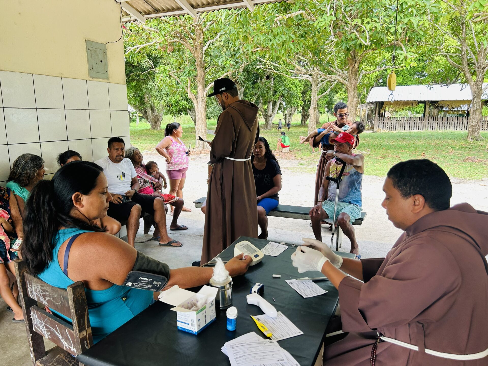 Mehrere indigene Menschen sitzen und stehen in einem offenen Unterstand, während ein Ordensmann in brauner Kutte einer Frau den Blutdruck misst. Ein weiterer Ordensmann in brauner Kutte steht in der Mitte und spricht mit den Wartenden. Die Szene zeigt eine einfache medizinische Versorgung in einer einem Indianerdorf im Amazonsagebiet.