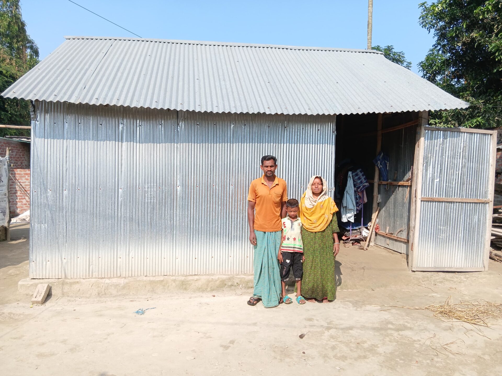 Eine Familie mit zwei Erwachsenen und einem Kind steht vor einem einfachen Haus aus Wellblech; die Tür ist offen, und einige Gegenstände sind im Inneren sichtbar.