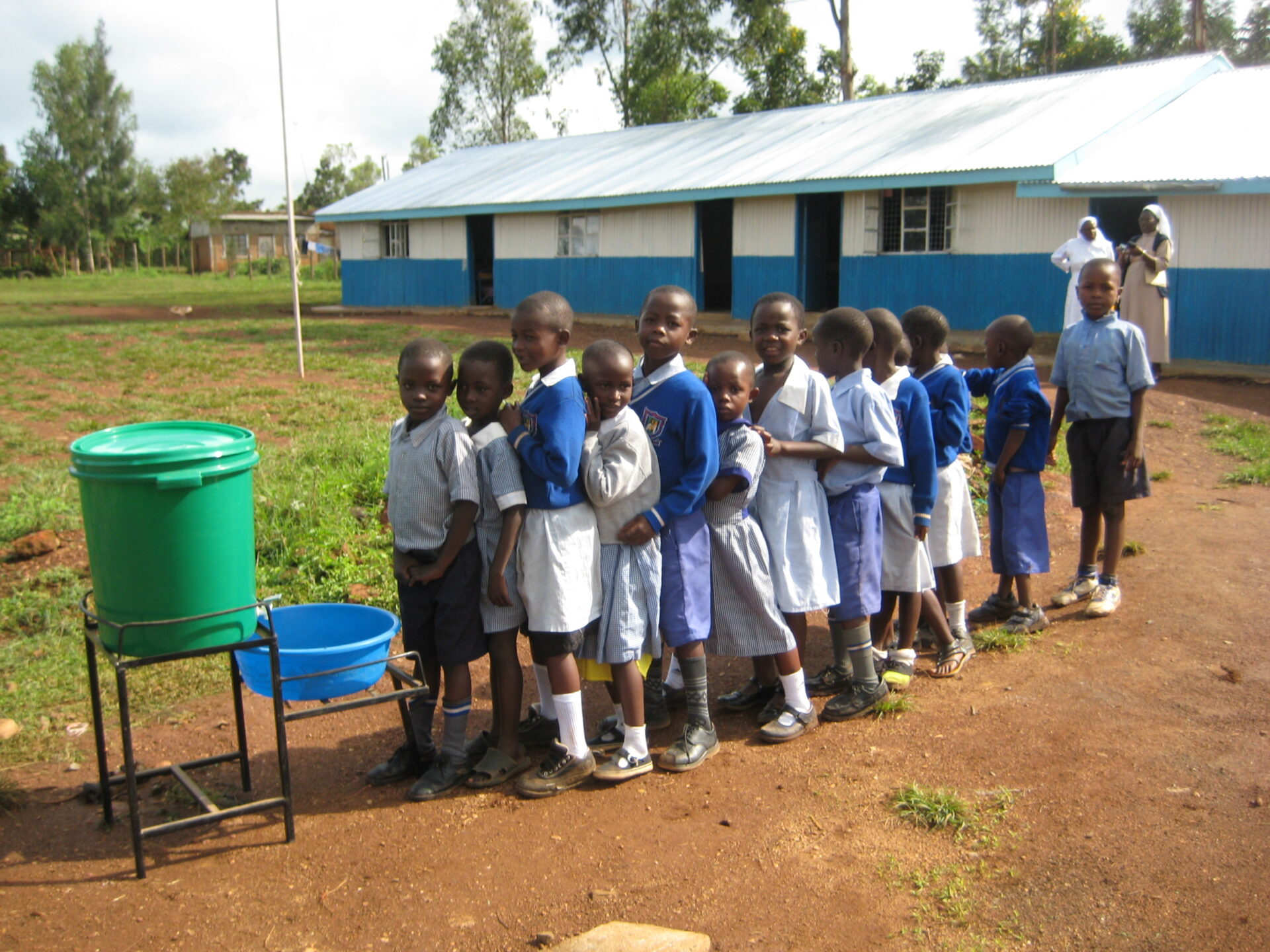 Eine Gruppe von Schulkindern in Uniform steht draußen in einer Reihe vor einer einfachen Handwaschstation; im Hintergrund ist ein langes Schulgebäude zu sehen, daneben stehen zwei Ordensfrauen, eine im weißen, die andere im beigen Habit.