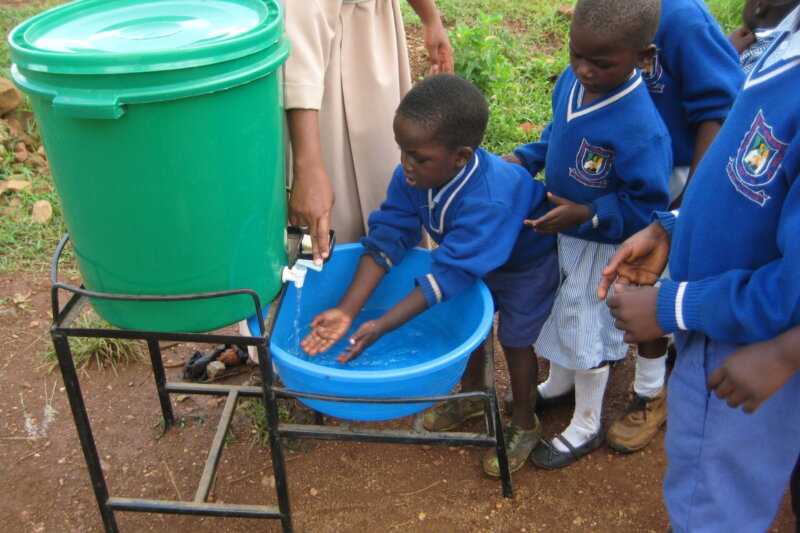Mehrere Kinder in Schuluniform stehen im Freien an einer einfachen Handwaschstation; ein Kind wäscht sich die Hände unter fließendem Wasser, während eine Ordensfrau im weißen Habit den Hahn betätigt und andere Kinder warten.