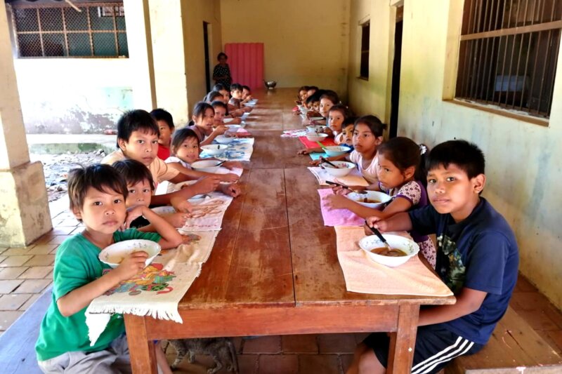 Viele Kinder sitzen an einem langen Holztisch unter einer Veranda und essen aus Schüsseln; die meisten schauen zur Kamera, während am Ende des Tisches eine erwachsene Person steht.