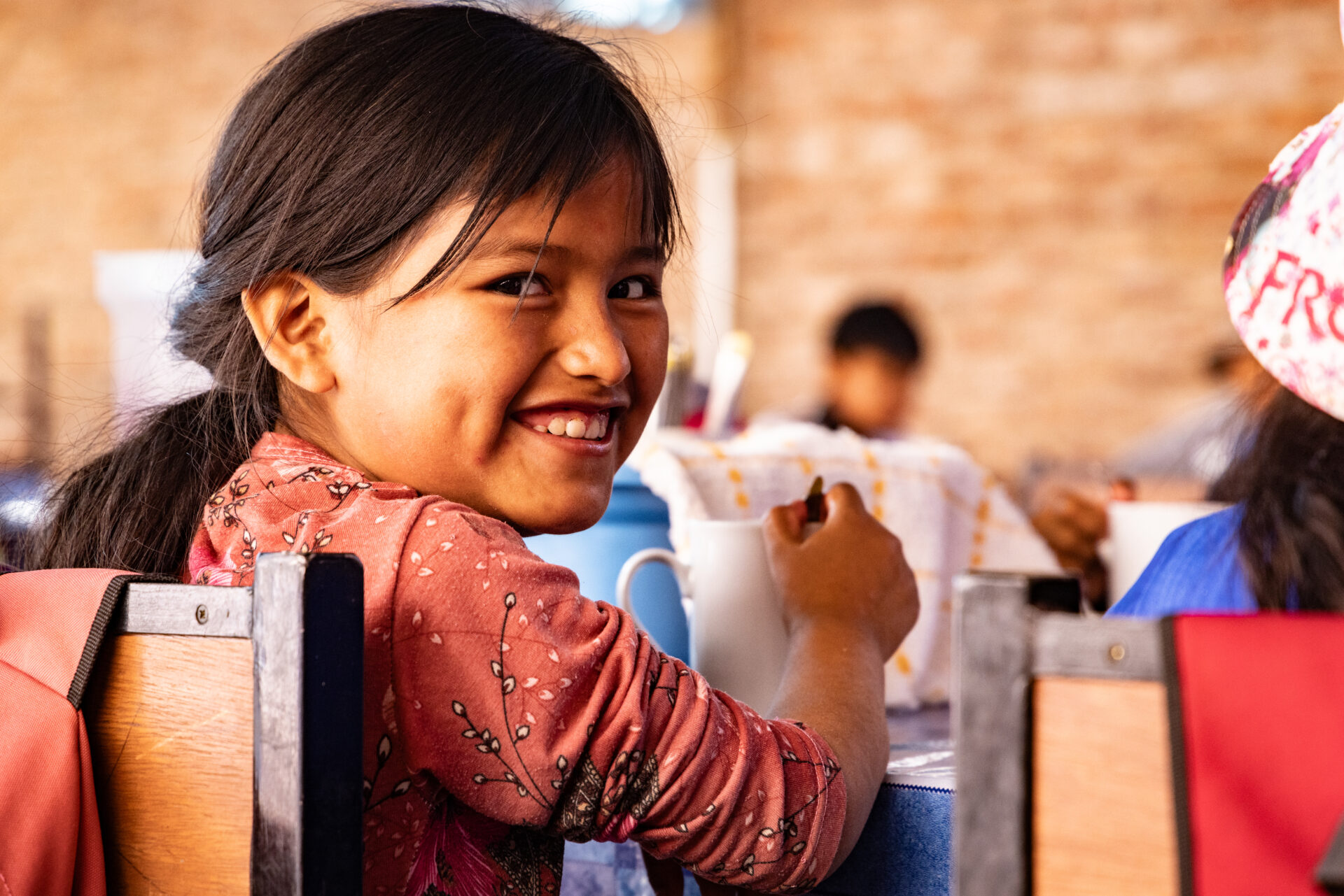 Ein lächelndes Mädchen sitzt an einem Tisch, dreht sich zur Kamera und hält eine Tasse in der Hand; im Hintergrund sind unscharf weitere Kinder in dem Raum zu sehen.