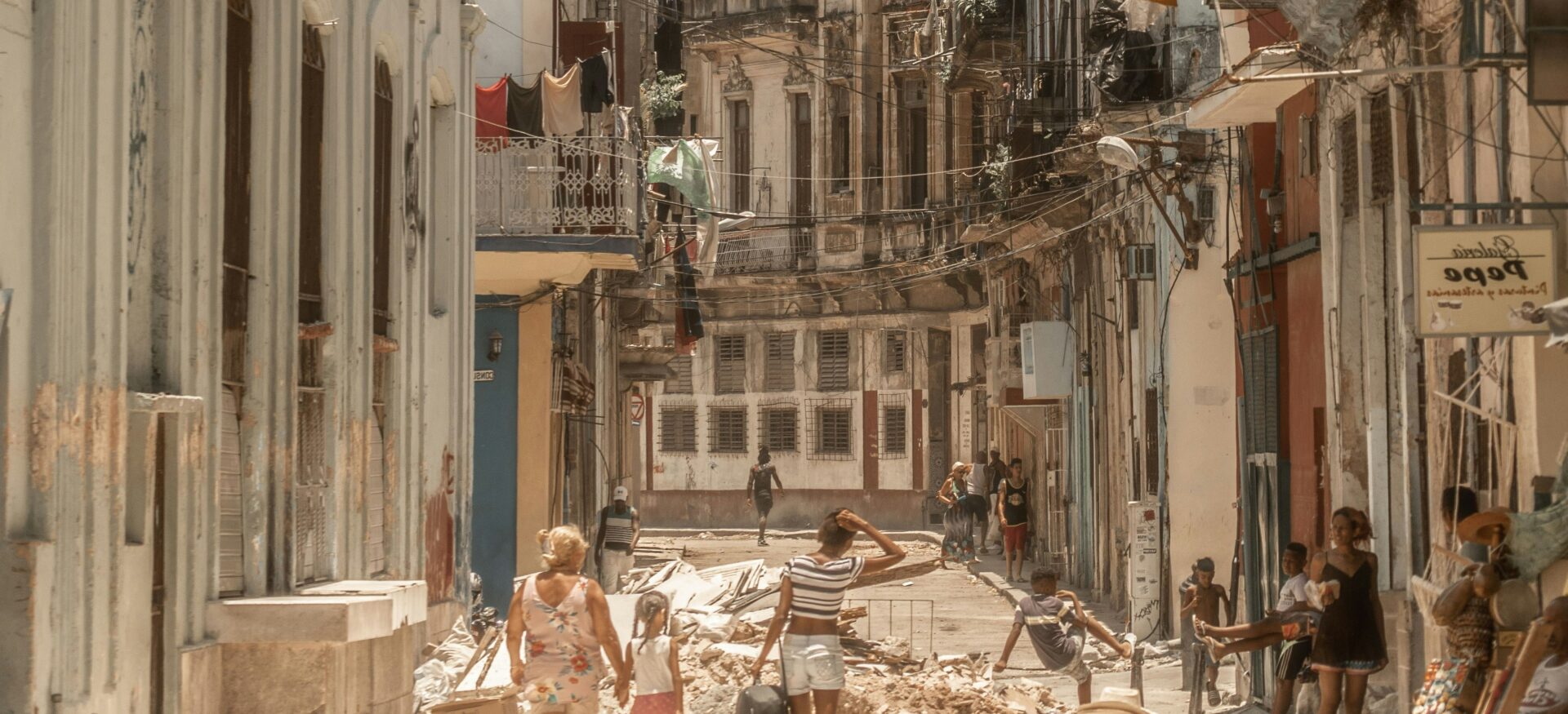 Eine enge Straße mit alten, teils beschädigten Häusern in Havanna. Zwischen Schutt und bröckelnden Fassaden bewegen sich Menschen durch die Straße, während über ihnen zahlreiche Stromkabel verlaufen.