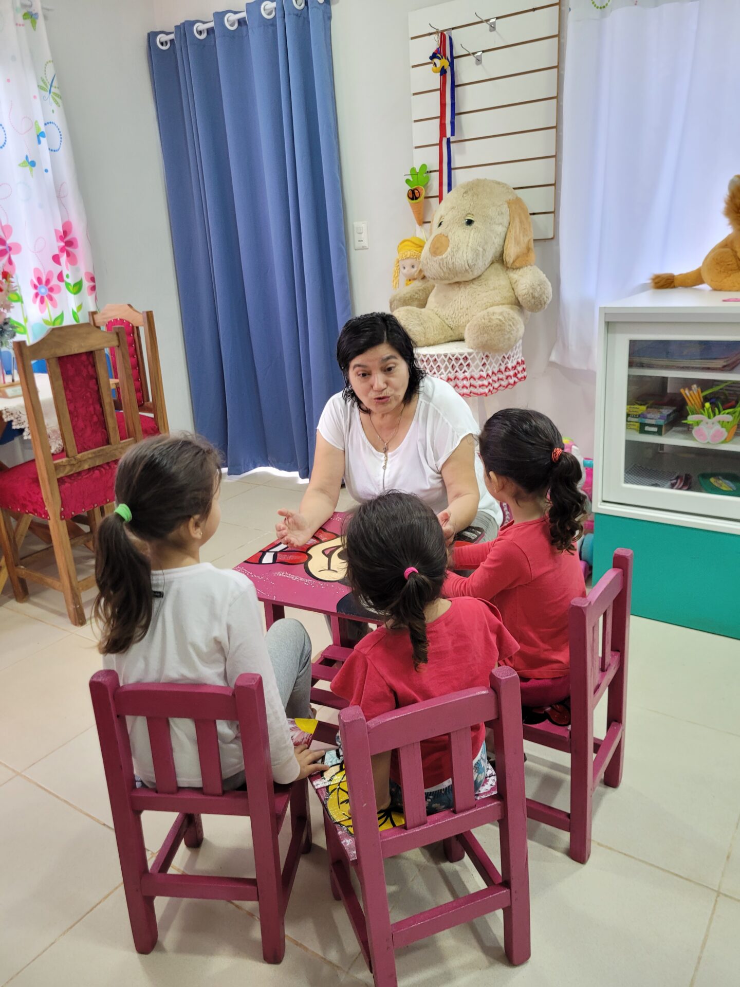 Eine Betreuerin sitzt mit drei kleinen Kindern an einem niedrigen Tisch in einem freundlich gestalteten Gruppenraum. Die Kinder hören aufmerksam zu, während die Frau mit ihnen spricht; Spielzeug und Stofftiere schaffen eine warme und geschützte Atmosphäre.
