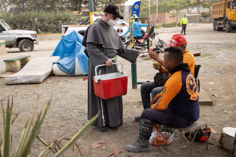 Franziskanerbruder verteilt Getränke an Mitarbeiter des Zivilschutzes nach dem Erdbeben in Venezuela. Im Hintergrund sind Fahrzeuge und weitere Einsatzkräfte zu sehen.
