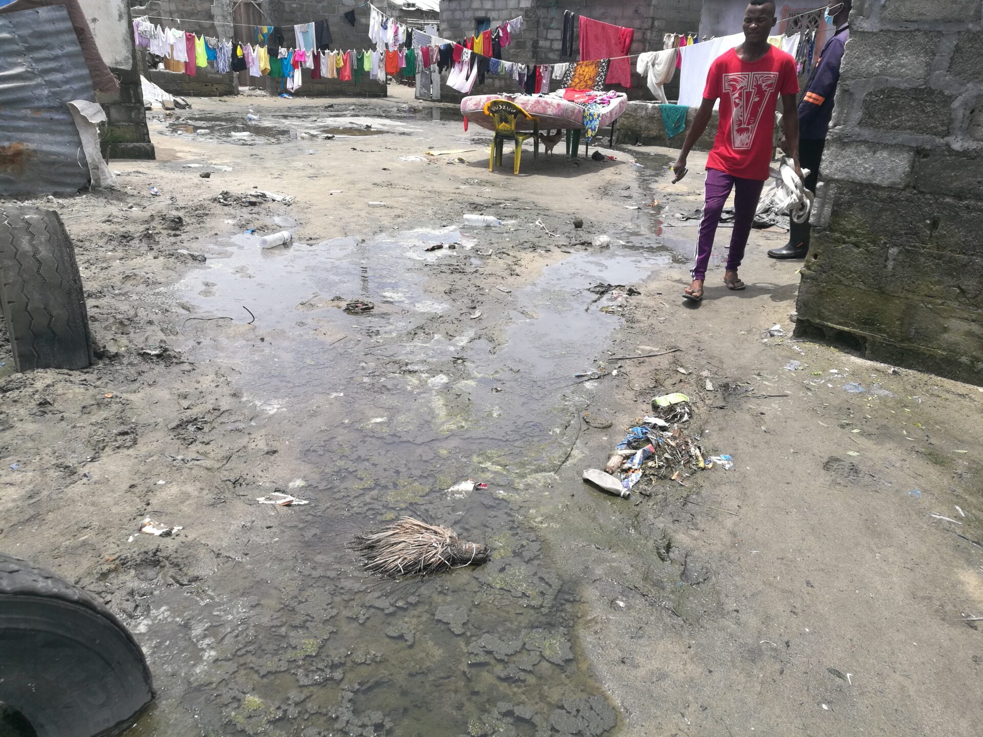 Eine verschmutzte Siedlung mit großen Wasserlachen, Schlamm und Abfällen zwischen einfachen Häusern. Im Hintergrund hängt Wäsche auf Leinen, während Menschen durch die von mangelhafter Infrastruktur geprägte Umgebung gehen.