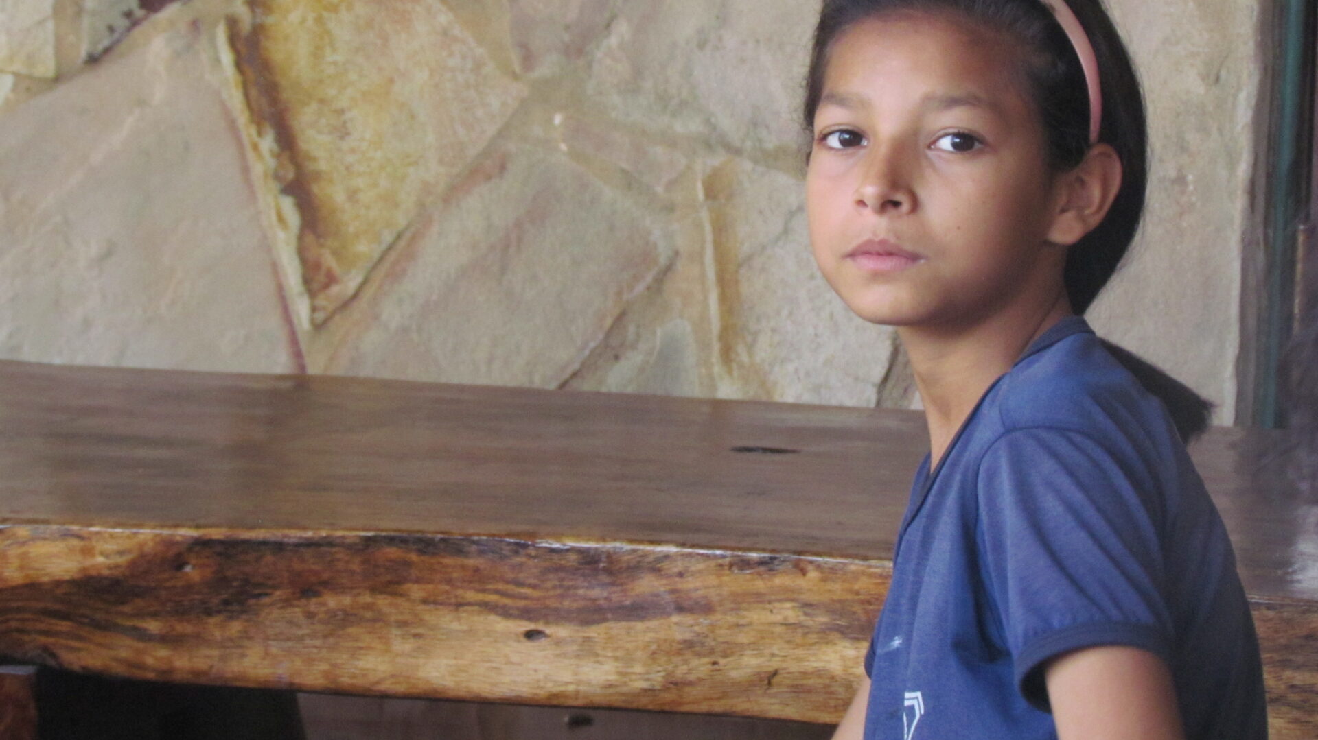 Ein Mädchen indigener Herkunft blickt ruhig in die Kamera. Es sitzt vor einer einfachen Steinwand und einem Holztisch, ihr Gesichtsausdruck wirkt nachdenklich und ernst.