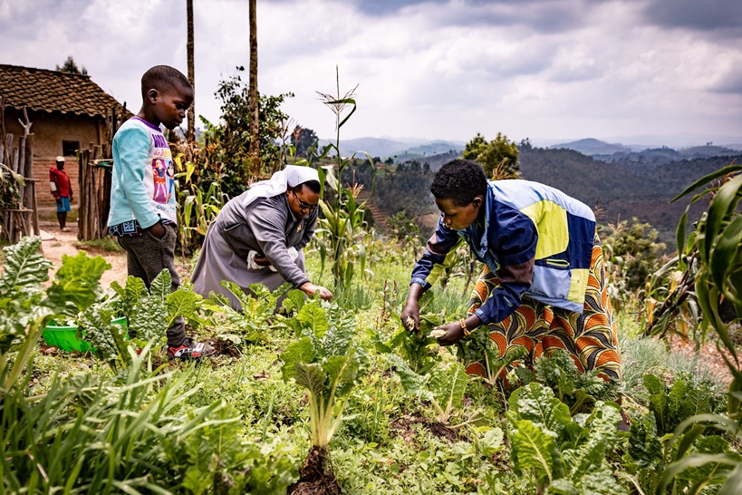 Eine Franziskanerschwester unterstützt gemeinsam mit einer Frau den Gemüseanbau in einem Dorf in Ruanda. Ein Kind steht daneben.