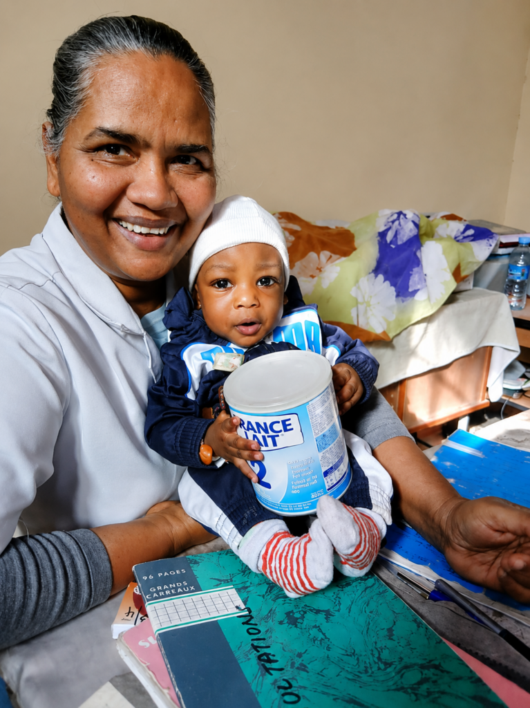 Eine lächelnde Frau hält ein Baby, das eine Dose Säuglingsnahrung in den Händen hält. Beide blicken in die Kamera und sitzen in einem einfach eingerichteten Raum.