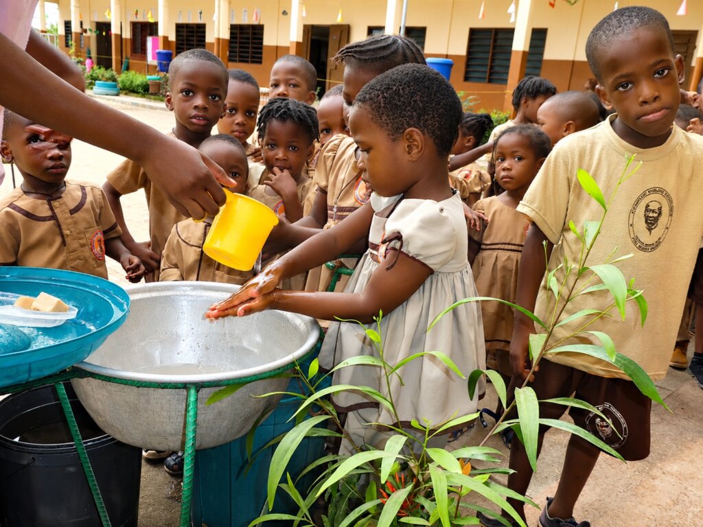 Mehrere Kinder stehen um ein großes Waschbecken im Schulhof. Ein Mädchen wäscht sich die Hände, während eine erwachsene Person Wasser aus einem gelben Becher darüber gießt. Die anderen Kinder schauen aufmerksam zu.