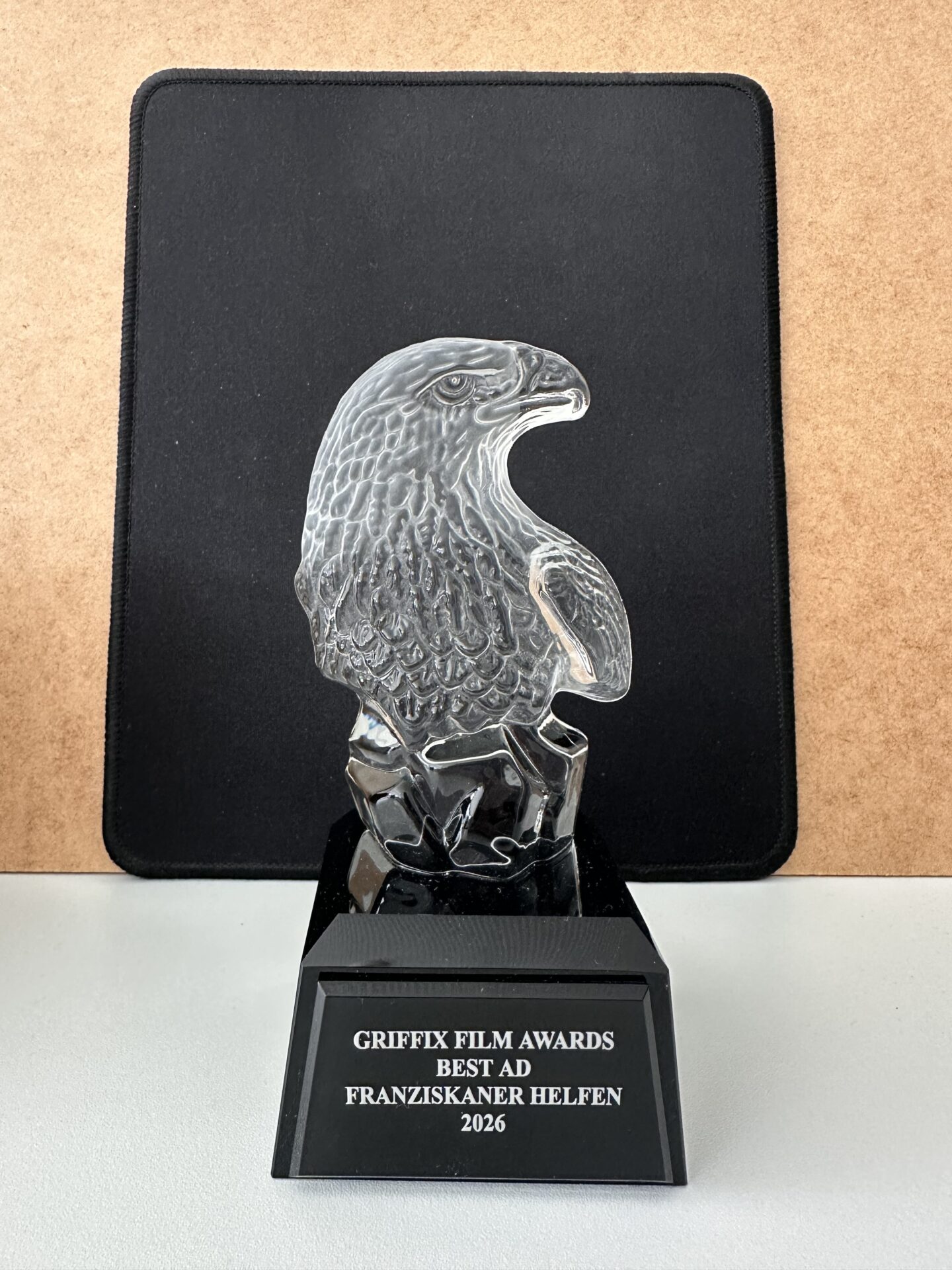 Gläserne Adler-Trophäe auf schwarzem Sockel mit der Aufschrift „Griffix Film Awards – Best Ad – Franziskaner Helfen – 2026“.