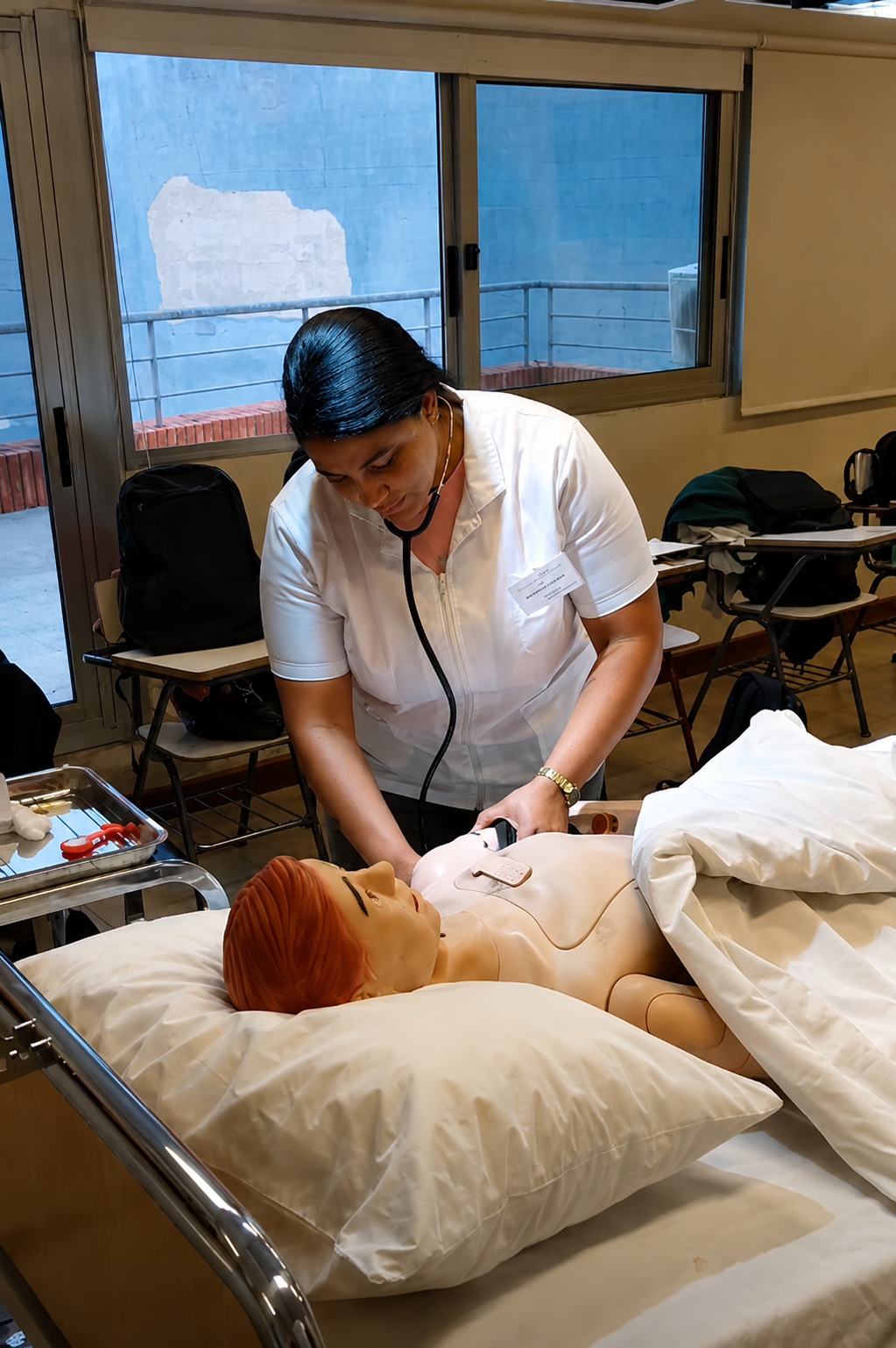 Eine medizinische Fachkraft in weißer Kleidung übt mit einem Stethoskop die Untersuchung einer Patientenpuppe, die in einem Krankenhausbett liegt.