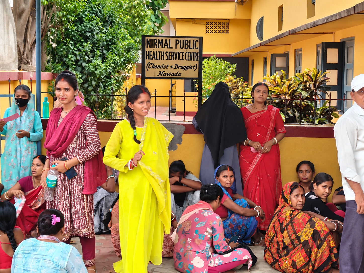 Mehrere Frauen in farbenfroher indischer Kleidung warten vor dem gelben Gebäude des Nirmal Public Health Service Centre auf medizinische Versorgung.