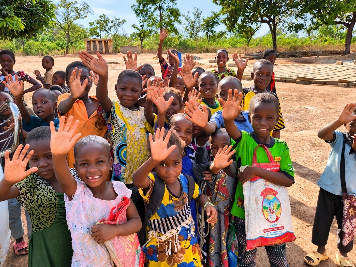 Eine große Gruppe von Kindern steht im Freien und winkt fröhlich in die Kamera. Viele lächeln und strecken ihre Hände in die Höhe. Im Hintergrund sind Bäume und ein einfaches Gebäude zu sehen.