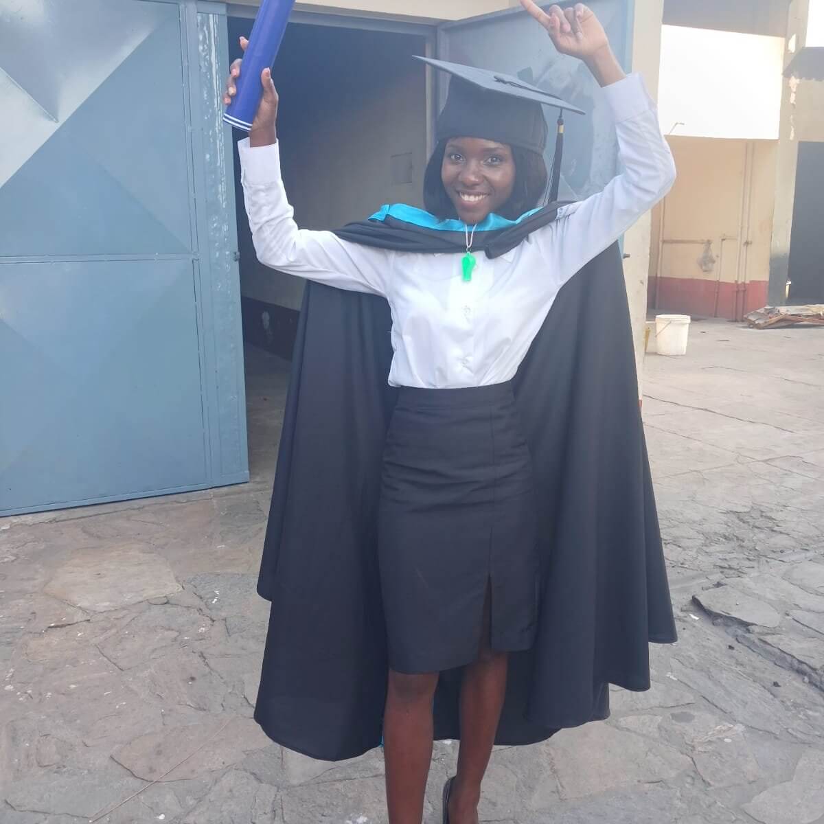 Junge Frau in schwarzem Talar und Doktorhut steht vor einem Gebäude, lächelt stolz in die Kamera und hält ihre Abschlussurkunde in die Höhe.