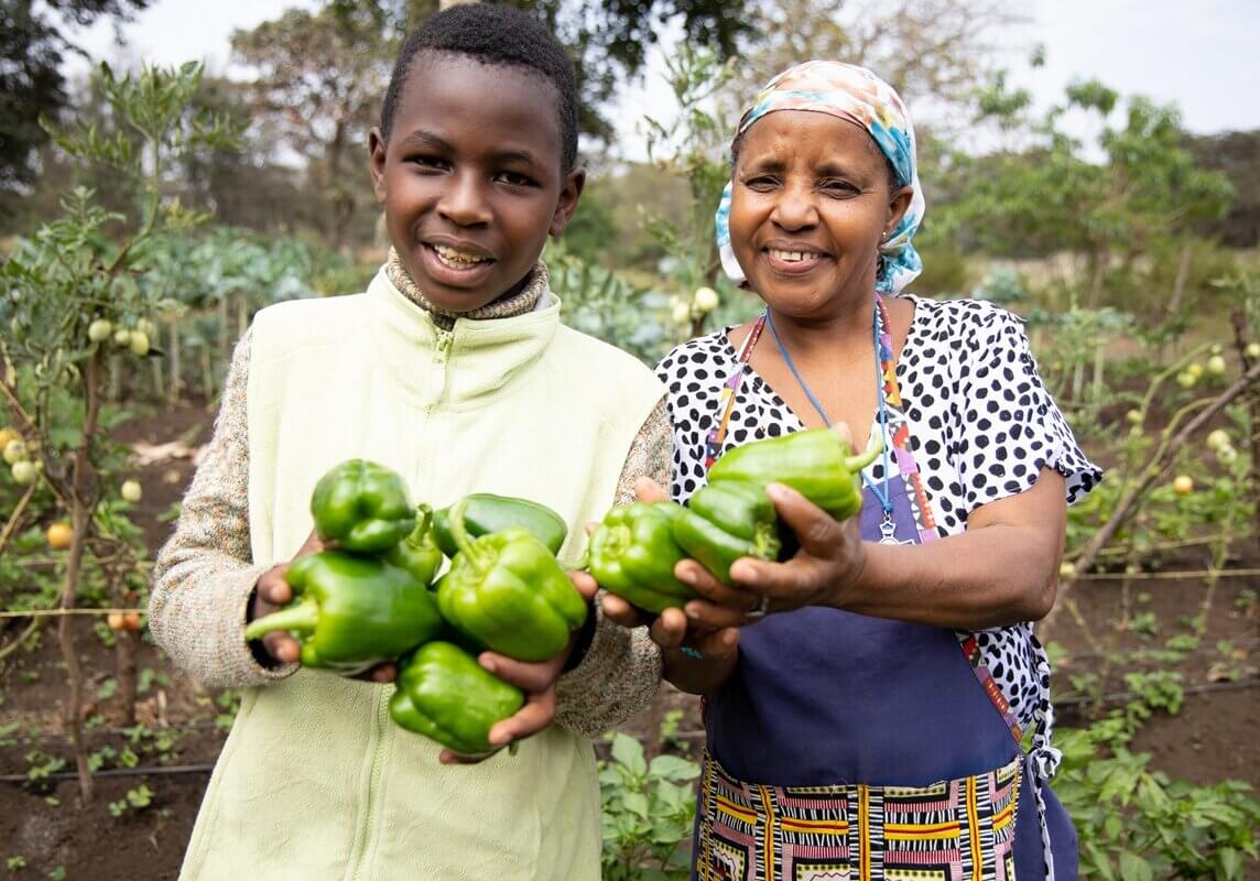 Ein Junge und eine Frau halten geerntete grüne Paprika in einem Gemüsegarten eines Projekts von Franziskaner Helfen in Tansania.