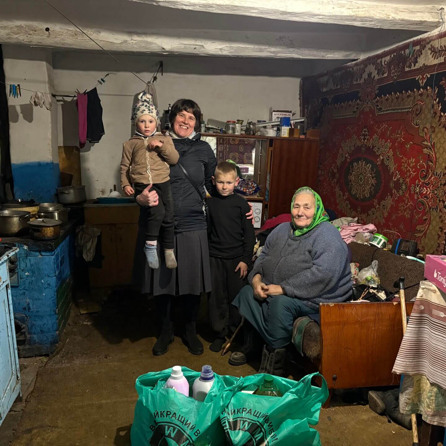 Eine Frau steht mit zwei Kindern in einem einfachen Wohnraum, daneben sitzt eine ältere Frau auf einem Sofa. Im Vordergrund stehen Taschen mit Hilfsgütern.