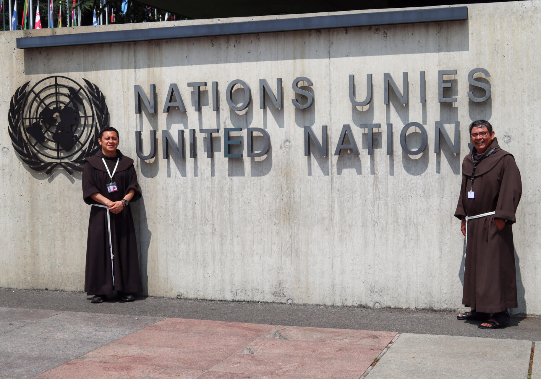 Zwei Franziskaner stehen vor dem Eingang der Vereinten Nationen neben dem UN-Emblem und dem Schriftzug „United Nations“.
