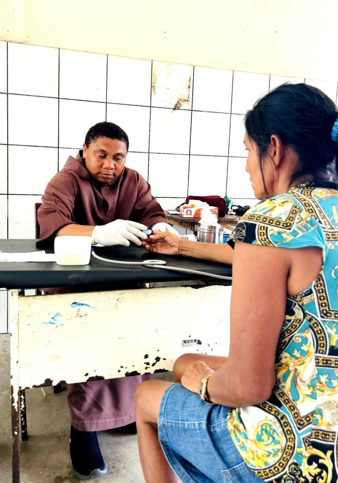 Ein Ordensmann in brauner Kutte sitzt an einem Tisch und misst einer Frau den Blutzucker an der Hand. Die Frau sitzt ihm gegenüber und streckt ihren Arm aus. Die Szene spielt in einem einfachen Raum und zeigt eine grundlegende medizinische Untersuchung.
