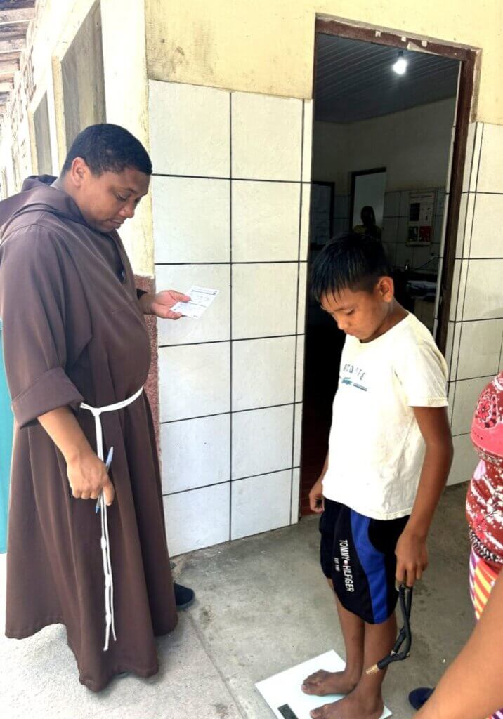 Ein Ordensmann in brauner Kutte steht neben einem Jungen, der barfuß auf einer Waage steht. Der Mann notiert Daten, während der Junge nach unten blickt.