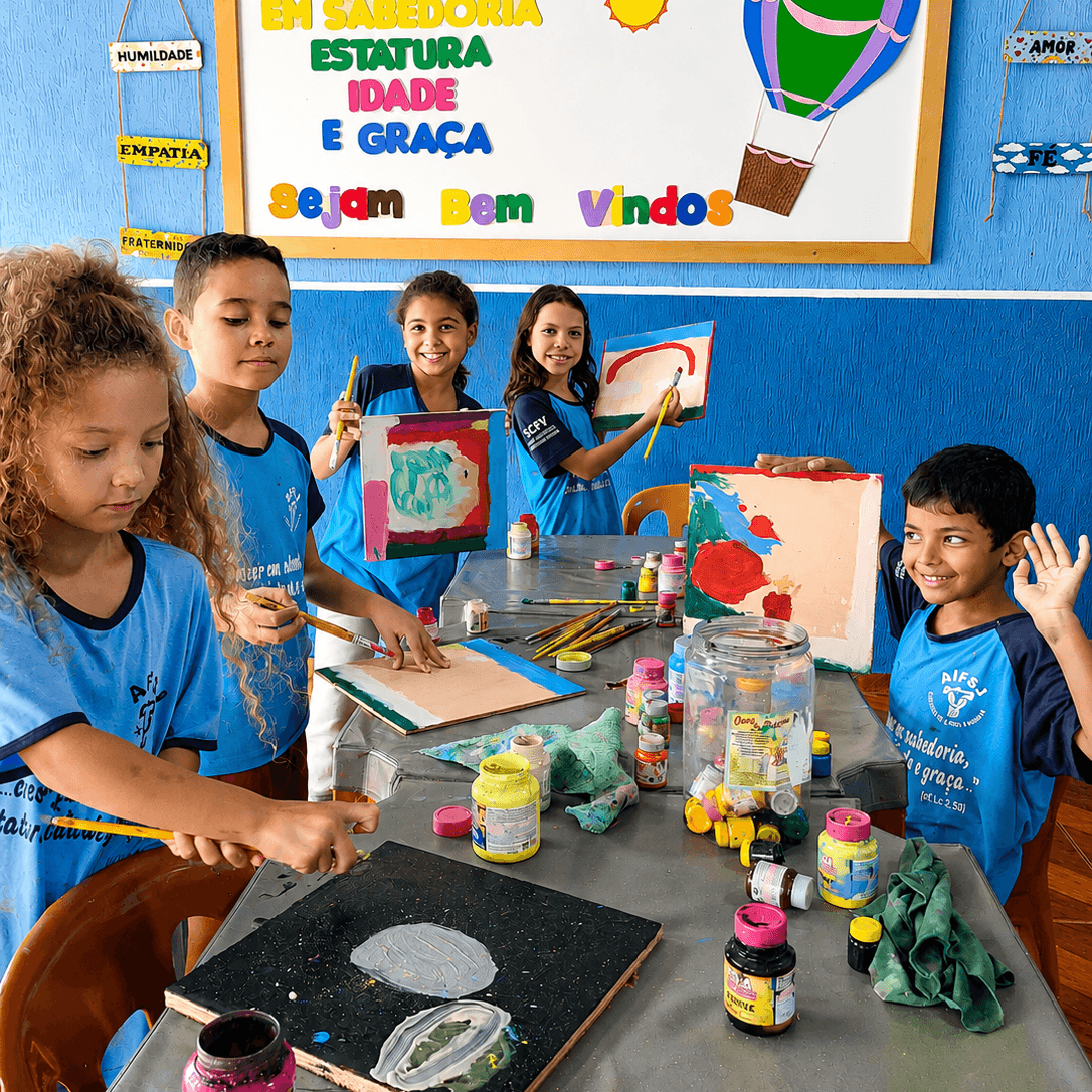 Sechs Kinder in blauen T-Shirts malen gemeinsam an einem großen Tisch. Einige präsentieren einige ihre bunten Bilder, während andere konzentriert mit Pinseln und Farben arbeiten.