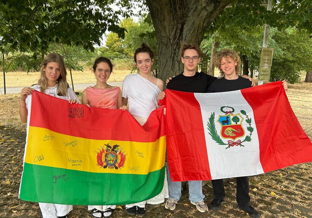 Fünf junge Freiwillige von Franziskaner Helfen stehen gemeinsam unter einem Baum und halten die Flaggen von Bolivien und Peru.