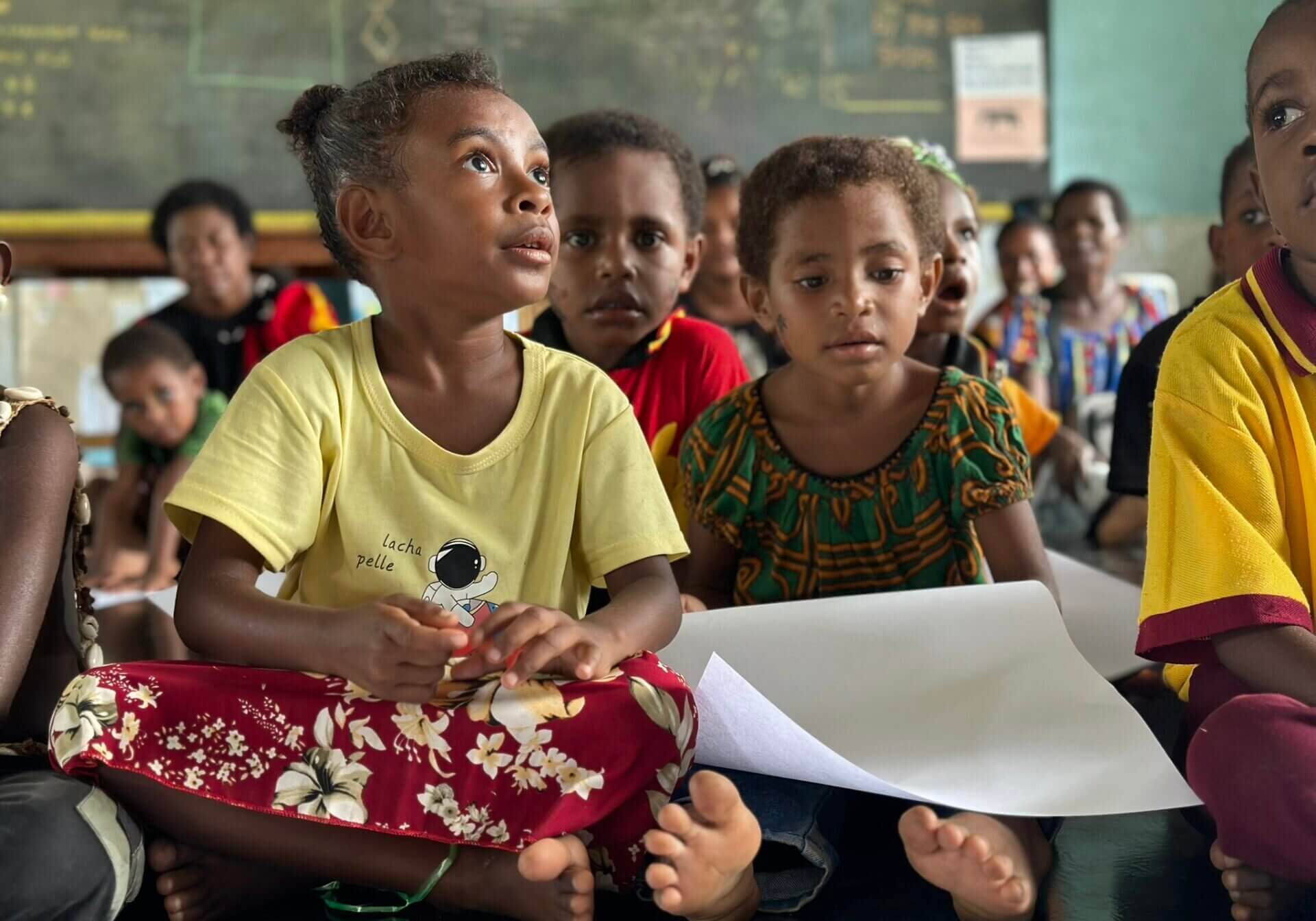 Barfüßige Kinder sitzen aufmerksam in einem Klassenraum; ein Mädchen hält ein Blatt Papier, im Hintergrund eine beschriebene Tafel.